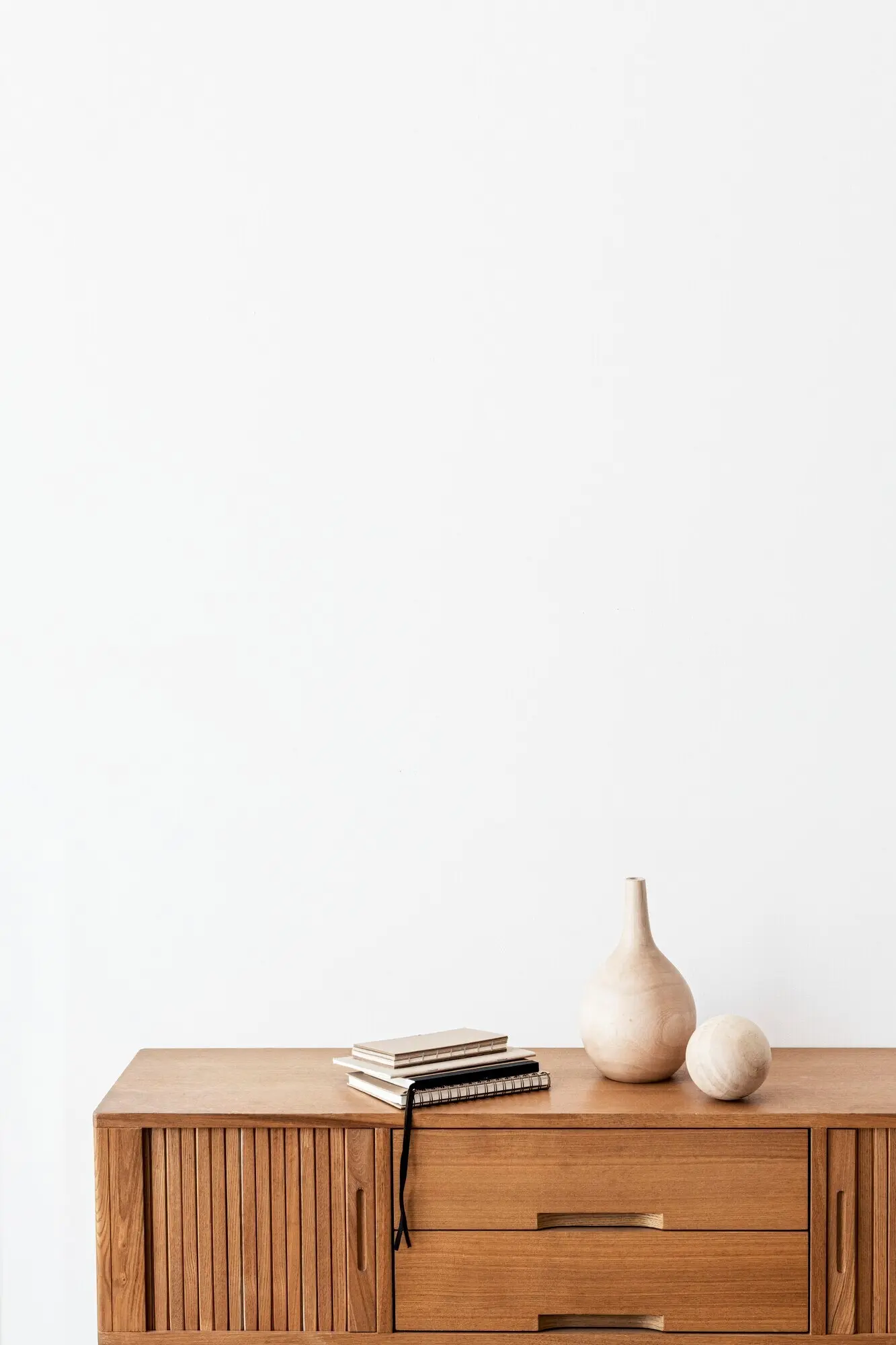 A stack of notebooks beside a wooden vase on a wooden cabinet in a white room.